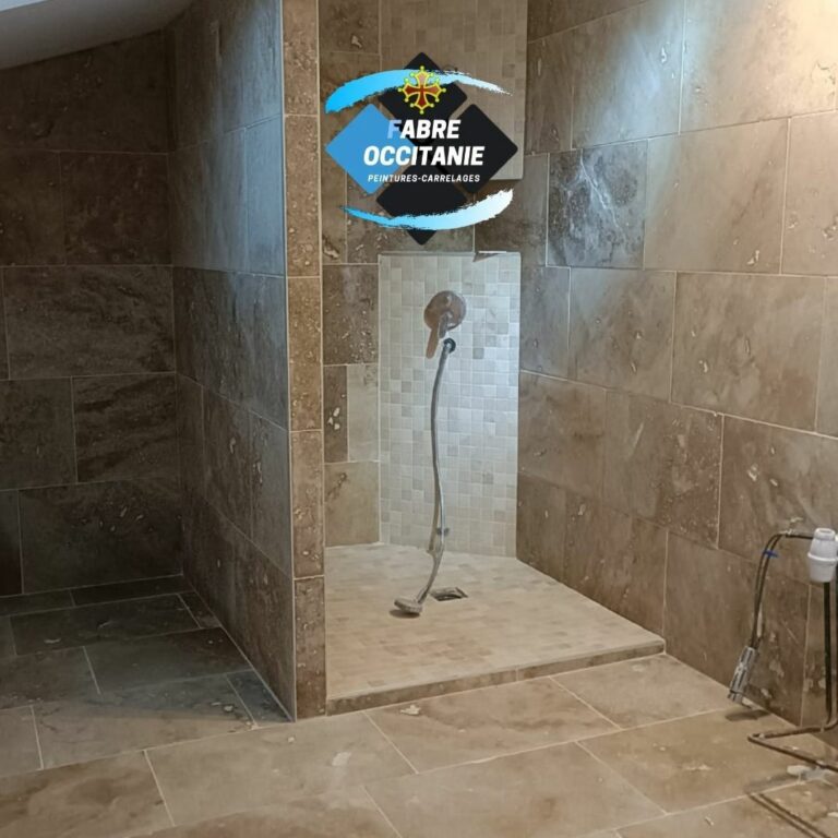 Fabre Occitanie - Une salle de bains moderne avec des murs et un sol carrelés beiges, avec un espace douche ouvert et une pomme de douche en acier inoxydable. un logo d'entreprise, "fabre occitanie", est superposé en haut.