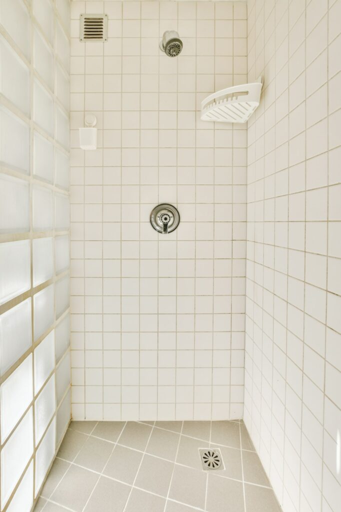 Fabre Occitanie - Un coin douche lumineux, propre et carrelé de blanc avec une pomme de douche Fabre Occitanie au plafond, des commandes de douche murales et une fenêtre en verre dépoli.