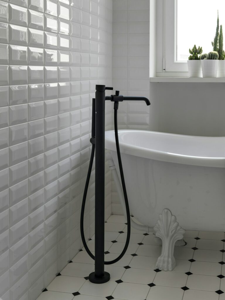 Fabre Occitanie - Une salle de bains moderne dotée d'un robinet noir autoportant à côté d'une baignoire Fabre Occitanie blanche avec des détails sur pattes, le tout contre un mur et un sol carrelés.