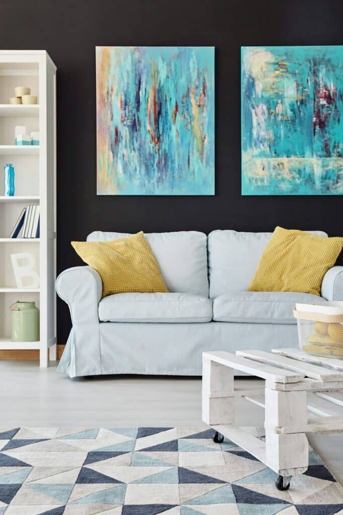 Fabre Occitanie - Salon moderne avec un canapé gris clair orné d'oreillers jaunes, des peintures bleues abstraites au mur et un tapis géométrique. Une table basse roulante blanche de Fabre Occitanie est au premier plan