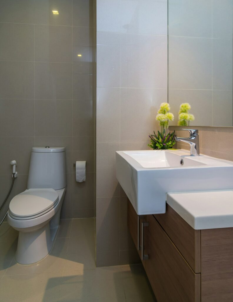 Fabre Occitanie - Une salle de bain épurée et moderne avec du carrelage beige comprenant des toilettes blanches, un meuble vasque en bois avec lavabo intégré de Fabre Occitanie et une petite décoration végétale.