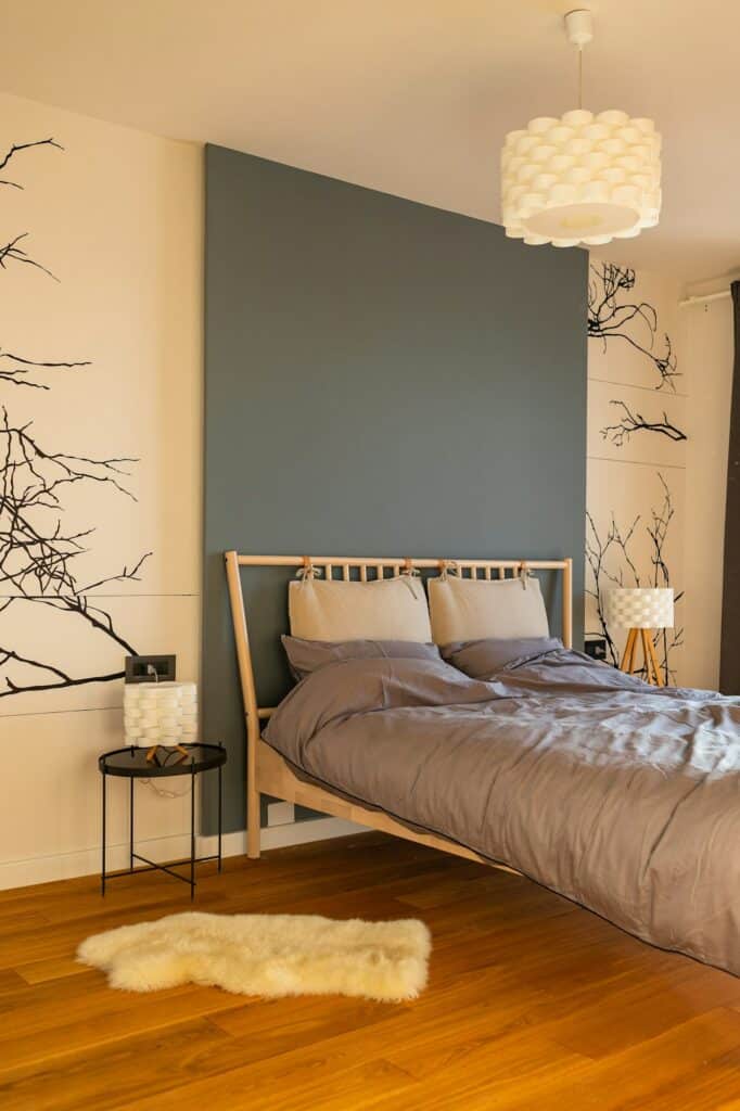 Fabre Occitanie - Une chambre moderne comprenant un lit gris avec des draps beiges, contre un mur d'accent bleu Fabre Occitanie, avec des motifs de branches décoratifs sur les murs adjacents et un tapis blanc moelleux sur le parquet.