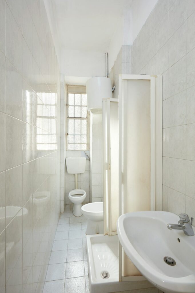 Fabre Occitanie - Intérieur de salle de bains étroit comprenant des toilettes blanches, un lavabo, une cabine de douche en verre dépoli et une petite fenêtre laissant entrer la lumière naturelle en Fabre Occitanie.