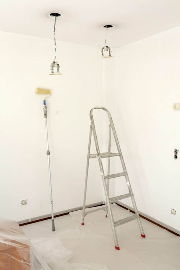 Fabre Occitanie - Une échelle et un rouleau à peinture dans une pièce en rénovation, aux murs blancs, aux prises électriques apparentes et au revêtement de sol de protection Fabre Occitanie.