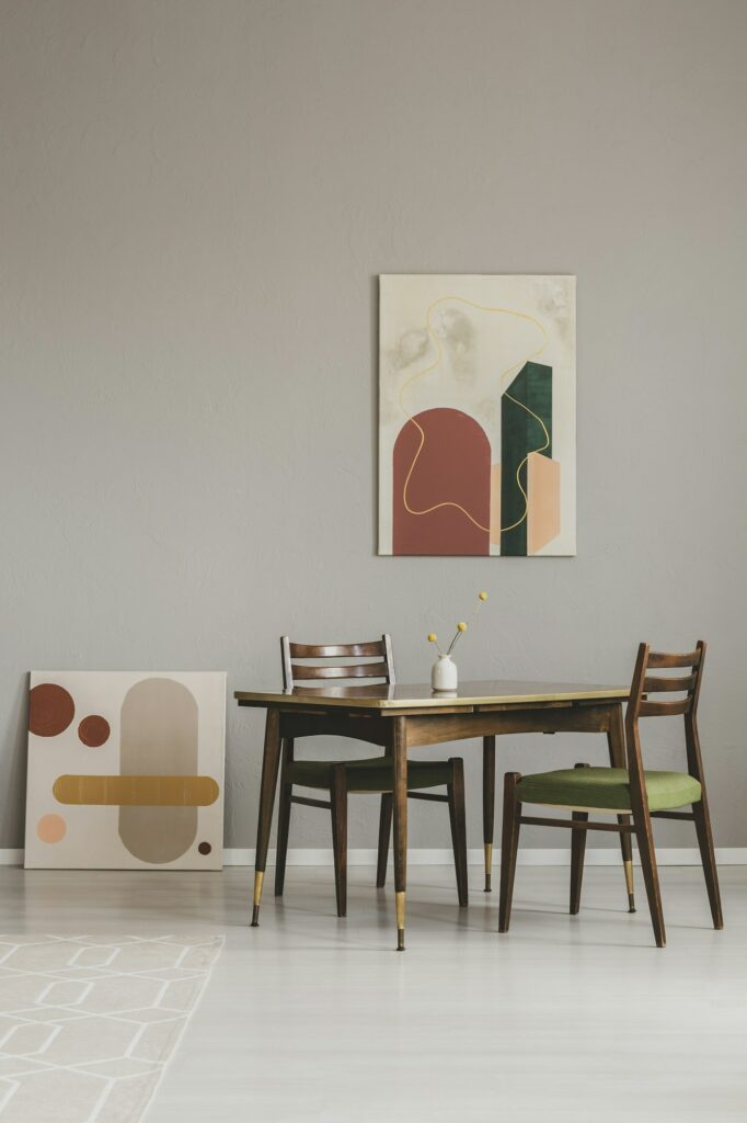 Fabre Occitanie - Un coin repas minimaliste avec une table en bois, quatre chaises et deux tableaux abstraits de Fabre Occitanie, l'un accroché au mur et l'autre adossé.