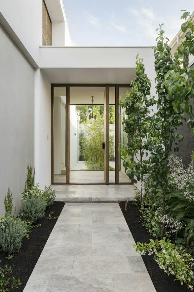Fabre Occitanie - Une entrée de maison moderne avec une porte en verre transparent encadrée de panneaux en bois, flanquée de plantes vertes luxuriantes et d'une allée en pierre grise à Fabre Occitanie.