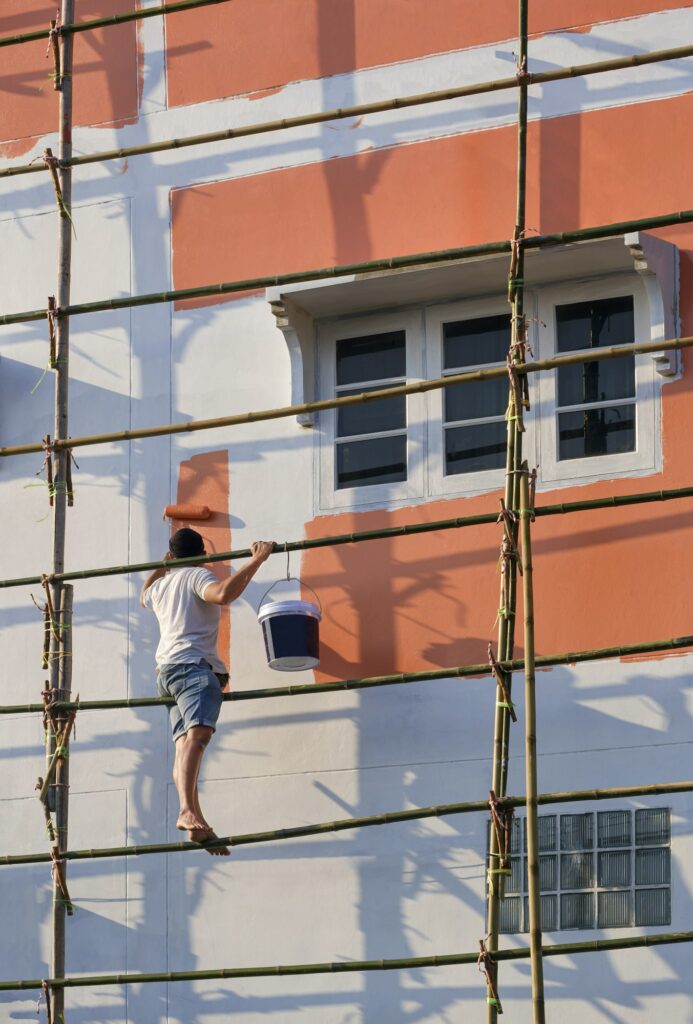 Fabre Occitanie - Une personne sur un échafaudage en bambou peint l’extérieur d’un bâtiment orange en Occitanie, en se concentrant sur une partie près d’une fenêtre.