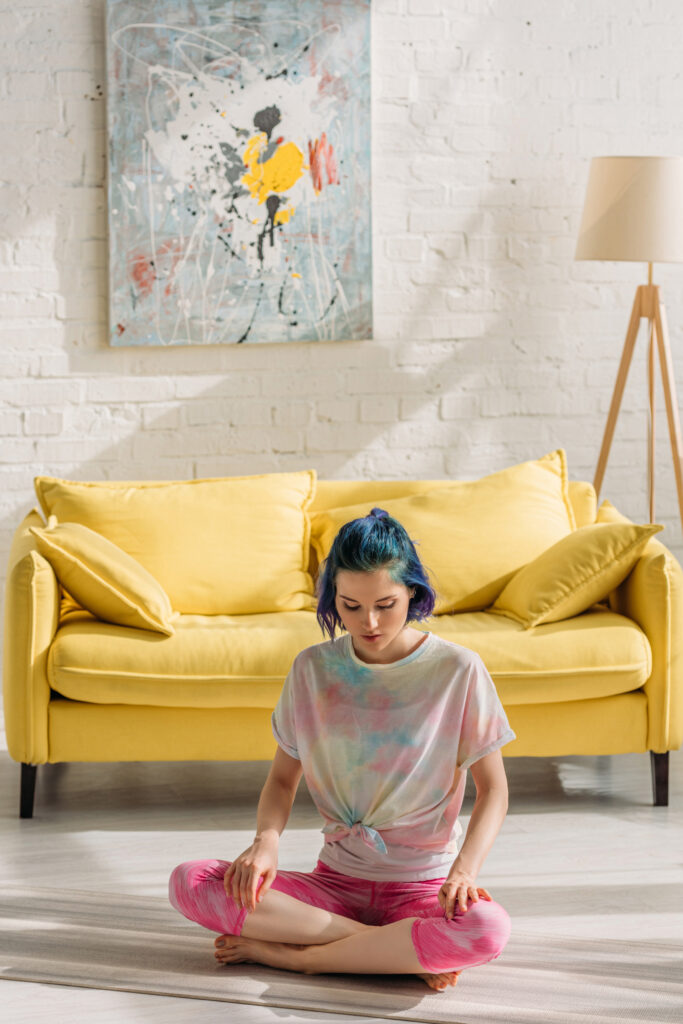 Fabre Occitanie - Une femme aux cheveux bleus médite par terre devant un canapé jaune et un tableau abstrait de Fabre Occitanie dans un salon lumineux.