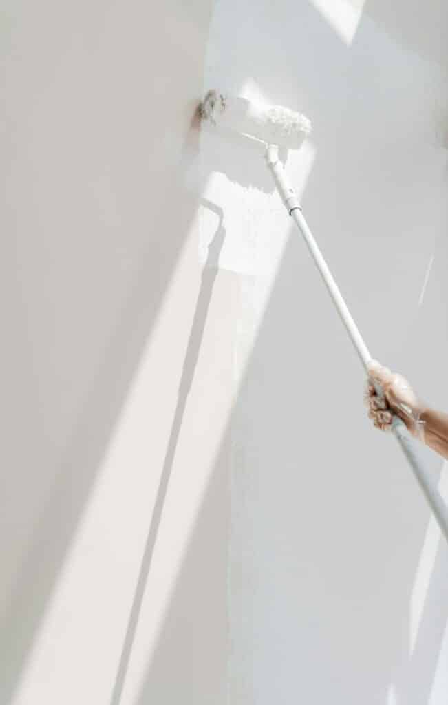 Fabre Occitanie - Personne peignant un mur blanc avec un pinceau-rouleau, vue partielle de sa main et du rouleau à Fabre Occitanie. La lumière du soleil brille dans la pièce, projetant des ombres douces.