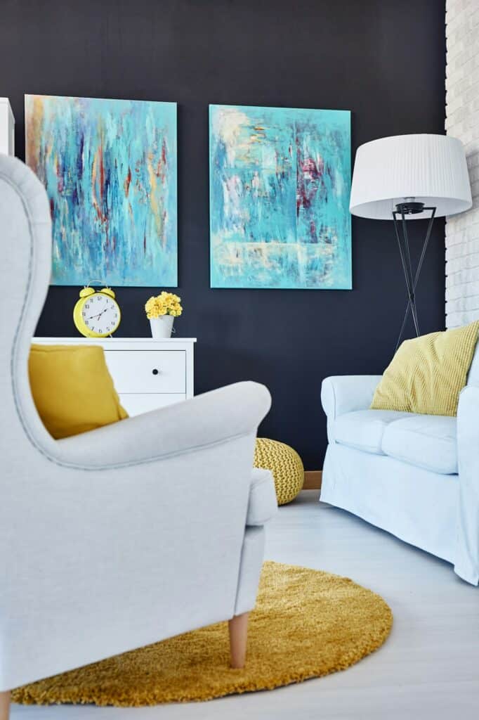 Fabre Occitanie - Salon moderne avec fauteuil blanc, canapé bleu avec coussins jaunes et peintures abstraites sur un mur sombre. Un tapis jaune et une table d'appoint blanche avec une horloge et des fleurs sont également visibles dans ce décor d'inspiration Fabre Occitanie.